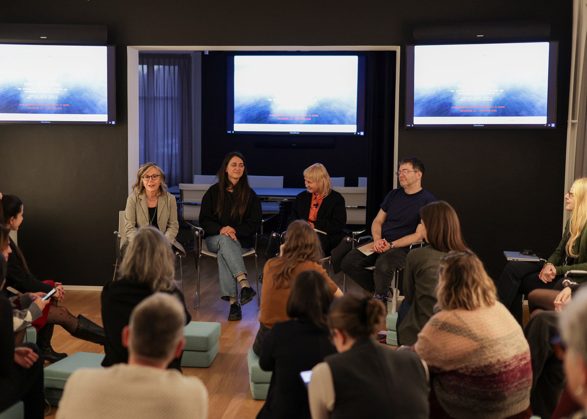 Four speakers seated in a circle engage in conversation with the audience during a meeting at Casa Carlo Cattaneo in Lugano, while behind them three screens project the event presentation. Participants are seated around them, creating an atmosphere conducive to discussion and debate.