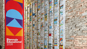 Banner verticale della Biennale Educational 2026 accanto a un'installazione composta da centinaia di disegni realizzati da bambini, appesi in lunghe colonne su una parete in mattoni all'interno di uno spazio espositivo