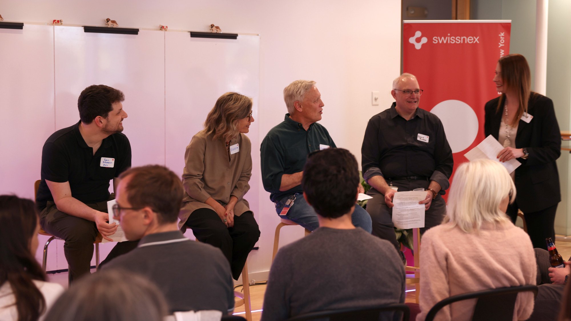 Un gruppo di persone sedute su sgabelli partecipa a una discussione in un ambiente informale, mentre una moderatrice in piedi interagisce con loro; sullo sfondo un roll-up Swissnex e una lavagna bianca.