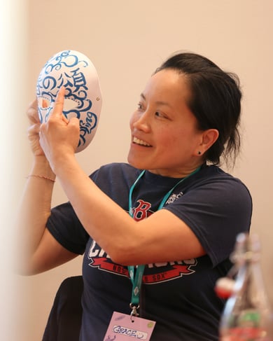 Una donna sorride mentre indica i dettagli di una maschera decorata che tiene in mano, durante un’attività di gruppo in un ambiente informale.