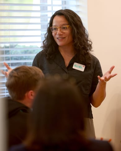 Una donna con occhiali parla al gruppo gesticolando con le mani, mentre i partecipanti ascoltano durante un incontro in un ambiente luminoso.