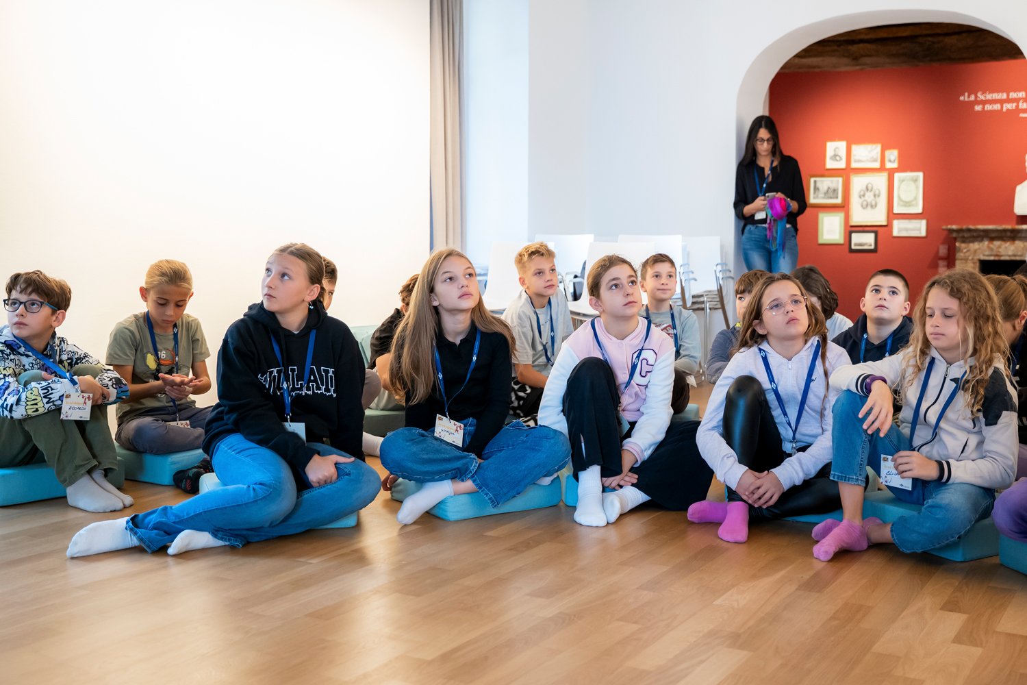 Bambini seduti a terra durante un laboratorio didattico, mentre ascoltano con attenzione in uno spazio museale.