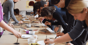 Un gruppo di persone chine su un lungo tavolo dipinge con pennelli e inchiostro su fogli di carta, partecipando a un laboratorio creativo collettivo.