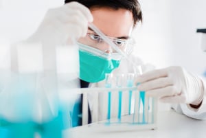 Ricercatore in laboratorio con mascherina, guanti e occhiali protettivi mentre utilizza una pipetta su provette contenenti liquido azzurro.