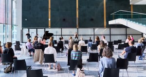 Ampia sala moderna con pareti scure e grandi vetrate. I partecipanti, seduti su sedie distanziate, assistono a un intervento su un palco essenziale accanto a un pianoforte. L’atmosfera è raccolta e riflessiva, tipica di un incontro culturale dedicato al dialogo e alla divulgazione.