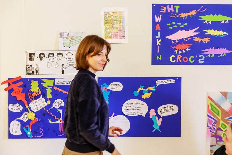 Woman with short brown hair walking past a wall decorated with colourful artistic posters. The posters include collages of abstract shapes, black-and-white comic strips, and text such as 'THE WALKIN CROCS' on a blue background, featuring stylised crocodile figures.