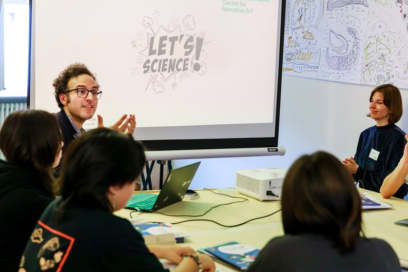 A physicist and an artist sitting around a table with students, explaining how the cartoonist managed to translate sounds and music onto paper. In the background, a screen displays the phrase "Let's Science."