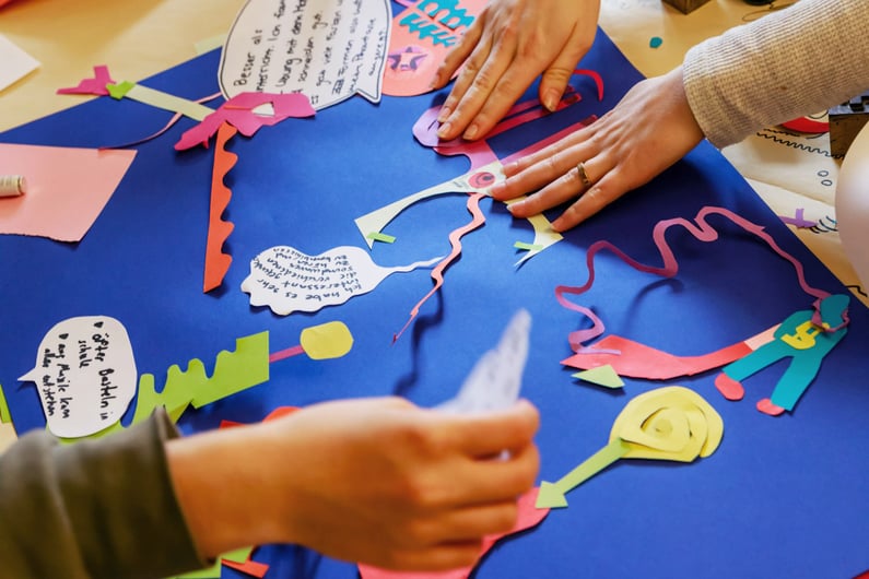 Hands of multiple people working on an artistic collage on blue cardboard, decorated with colourful paper shapes, arrows, stylised figures, and speech bubbles with handwritten text.