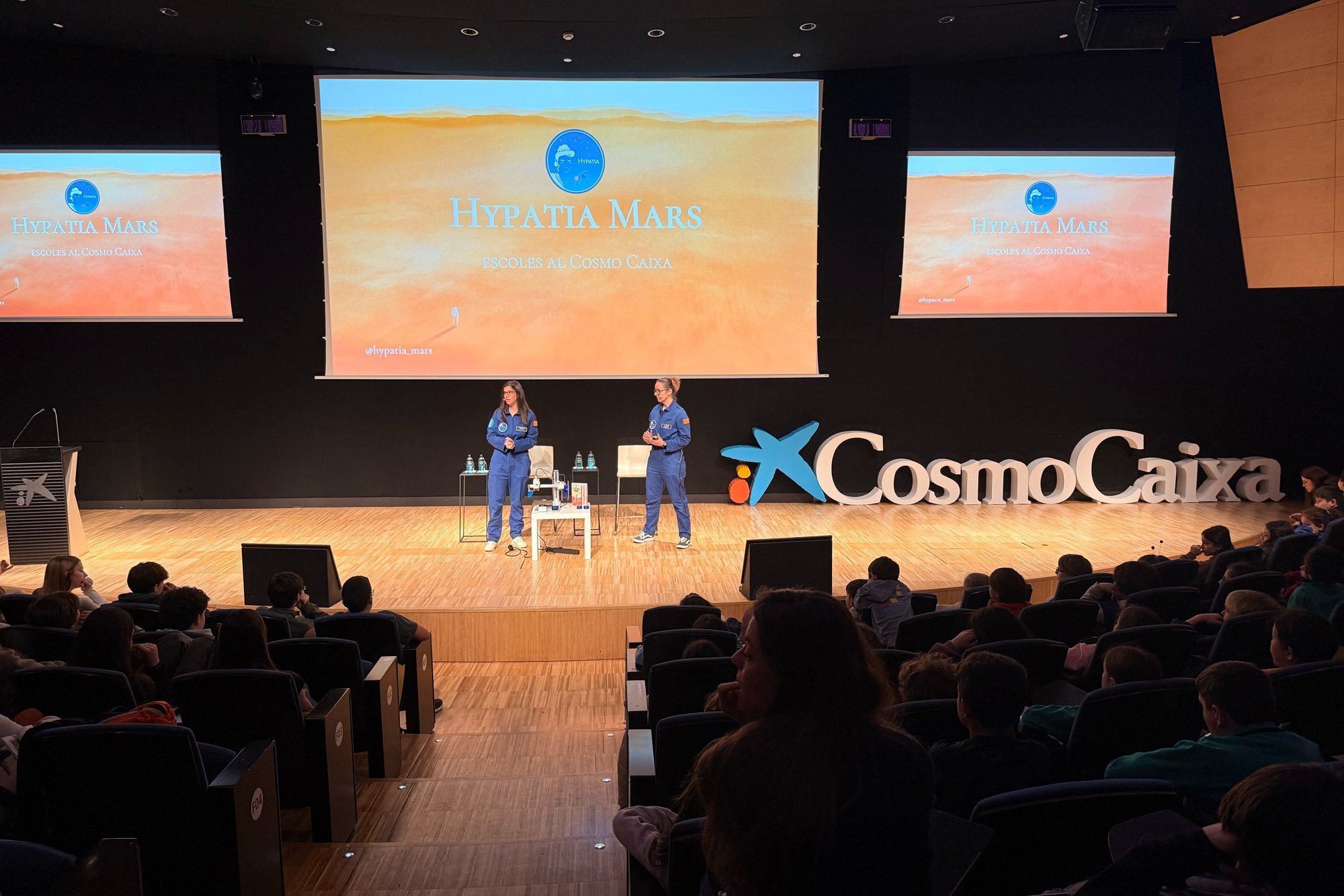 Vista ampia della sala del CosmoCaixa completamente occupata da studenti. Sul palco, le relatrici illustrano le missioni Hypatia, con slide che mostrano immagini di team e loghi delle missioni. L’atmosfera è coinvolgente e partecipata.