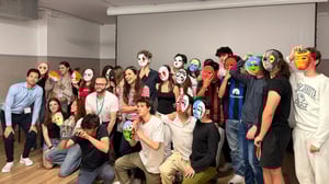 Gruppo numeroso di studenti e facilitatori durante un laboratorio HappyLab di IBSA Foundation a Barcellona, posano insieme indossando e mostrando maschere artistiche colorate realizzate durante l’attività.