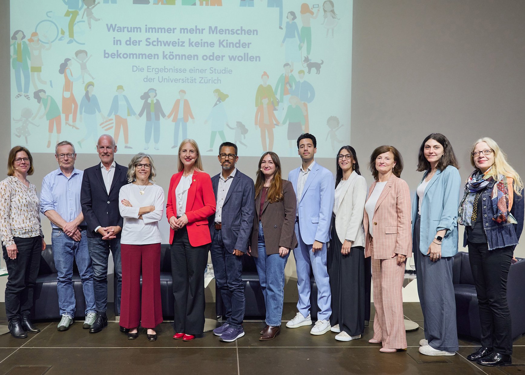 A group of speakers, organisers and participants posing in front of the poster for the event on parenting in Switzerland.