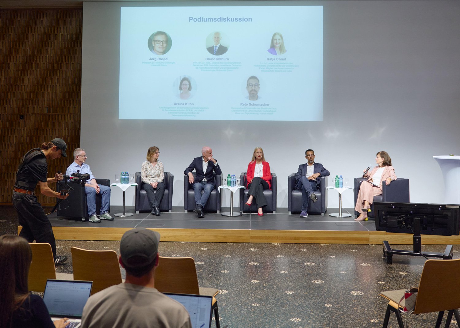 A wide-angle shot of the round-table discussion, with speakers on stage, the audience in the hall and videographers at work.