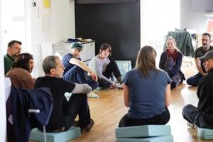 Gruppo di persone sedute in cerchio su cuscini azzurri all’interno di una sala moderna e luminosa, impegnate in una discussione collettiva.