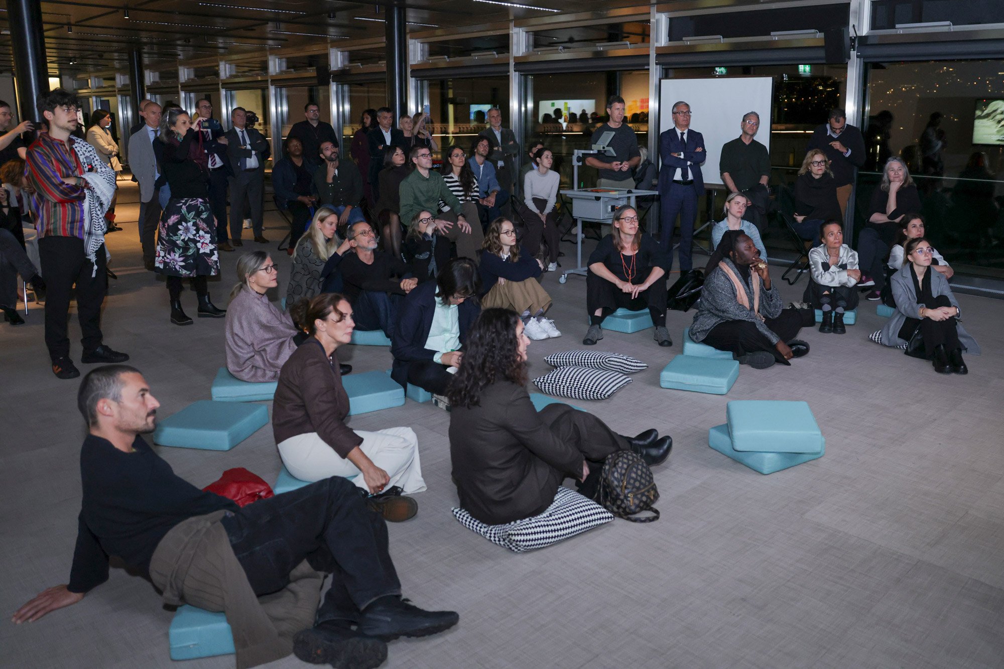 Pubblico seduto su cuscini azzurri e a terra durante un incontro serale in uno spazio museale a Lugano; i partecipanti ascoltano con attenzione una presentazione fuori campo, circondati da vetrate e da un allestimento contemporaneo.