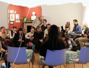 Partecipanti seduti in cerchio su cuscini durante un incontro in uno spazio storico con parete rossa, camino e busto in gesso; sullo sfondo una citazione sulla scienza, mentre alcune persone consultano il telefono in un momento di pausa o preparazione.