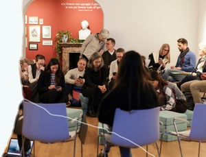 Participants seated in a circle on cushions during a meeting in a historic space with red walls, a fireplace, and a plaster bust; in the background, a quote about science, while some people check their phones during a break or preparation time